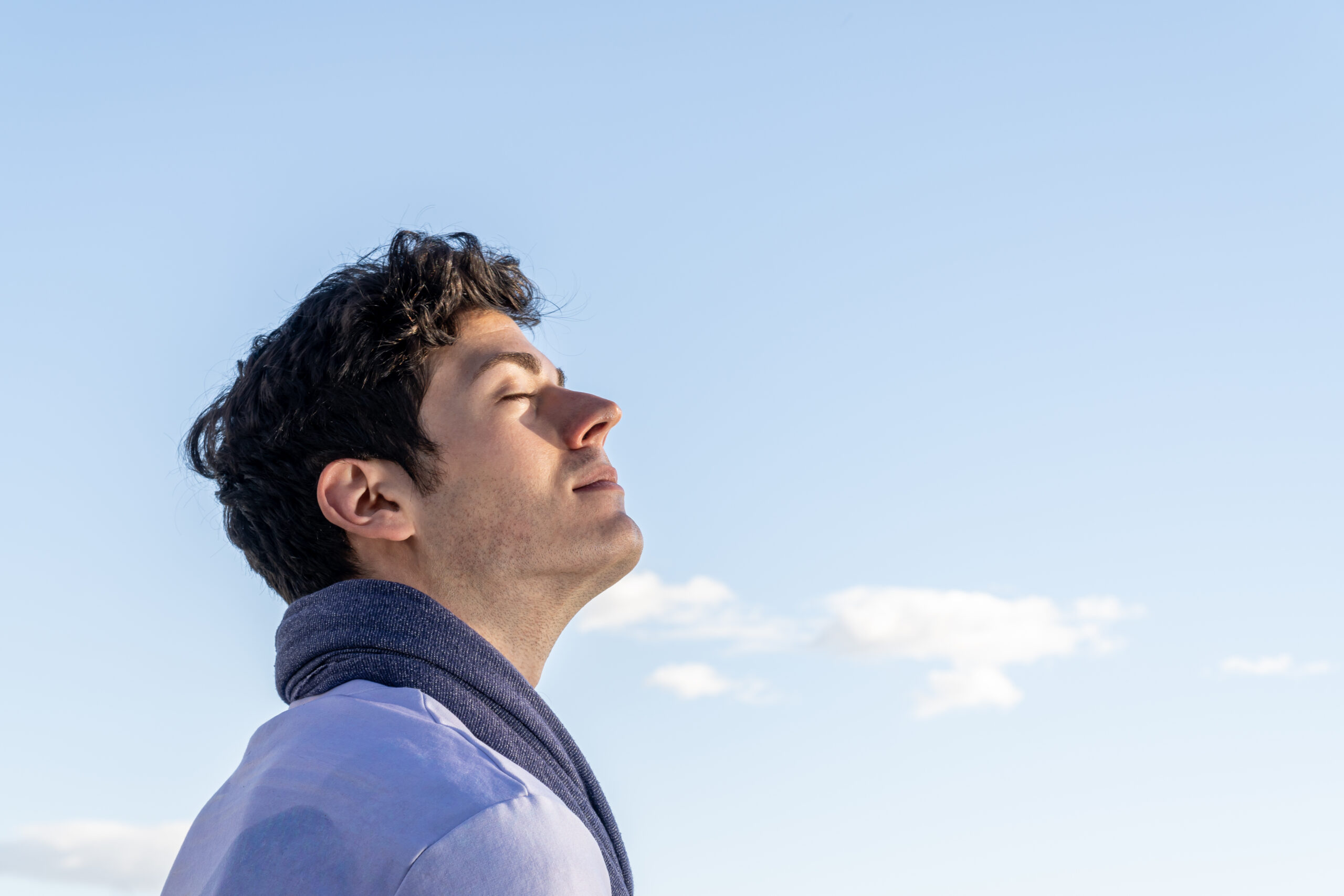 young boy with his head to the sky and eyes closed on a clear day woman sitting at the seashore